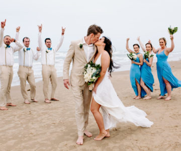 a bride and groom kissing on the beach with their bridal party